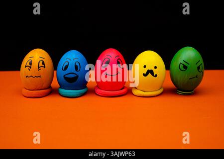 A row of brightly colored Easter eggs painted with playful emoji faces, placed on an orange surface against a black background. A fun and creative Eas Stock Photo