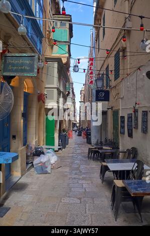Valletta, Malta - 01. October 2024: Charming narrow street lined with colorful buildings, string lights, and outdoor restaurant seating in historic Va Stock Photo