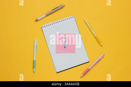 Notepad with pens and memo piece of paper with question mark on yellow background. Top view Stock Photo