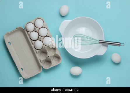 Cooking concept. Empty white bowl with whisk, egg tray on blue studio background. Top view. Stock Photo