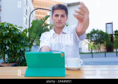 Smiling Caucasian man showing thumbs down while using tablet at outdoor cafe looking at camera Stock Photo