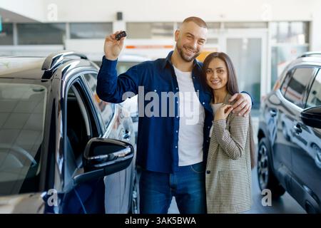 Smiling millennial couple holding car keys proudly in modern dealership showroom Stock Photo