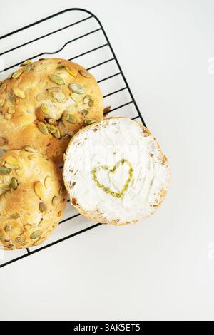 Delicious pumpkin seed bagels with cream cheese, topped with a heart-shaped pesto design, freshly baked on a cooling rack. Stock Photo