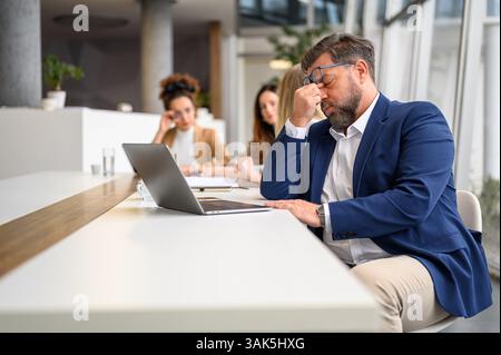 Stressed businessman rubbing his eyes while working over laptop with females colleagues in the background Stock Photo
