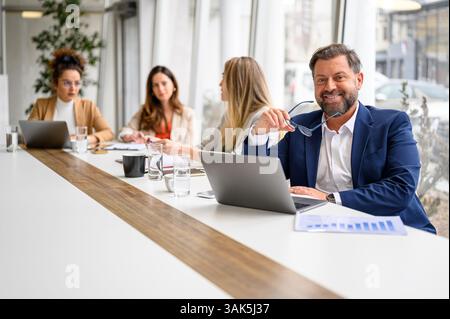 Businessman holding glasses and analyzing reports over laptop while sitting by female professionals in board room Stock Photo