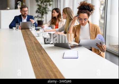 Focused accountant with laptop reviewing reports while teammates collaborating in the background in boardroom Stock Photo
