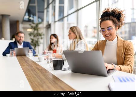 Smiling lawyer reviewing report on her laptop with legal colleagues collaborating in the background during professional meeting Stock Photo