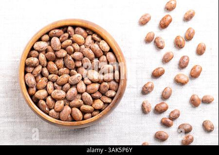 Pinto beans in a wooden bowl on linen fabric. Dried variety of common beans, Phaseolus vulgaris. Type of bean that are tan with reddish-brown specks. Stock Photo