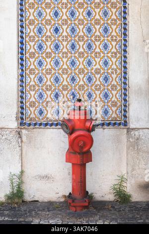 A weathered red fire hydrant stands on a cobblestone sidewalk in Lisbon, Portugal, against a rustic wall adorned with decorative Moorish tiles, captur Stock Photo