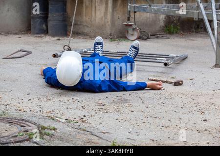 Construction worker wearing blue uniform and white safety helmet lying unconscious on the ground after a workplace accident with tools scattered nearb Stock Photo