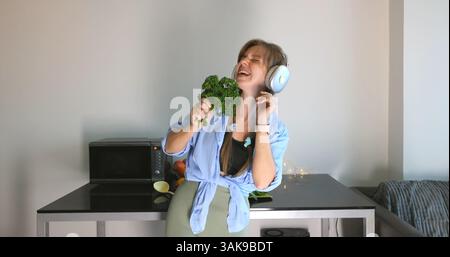 Woman in kitchen singing with kale, wearing headphones, joyful cooking Stock Photo