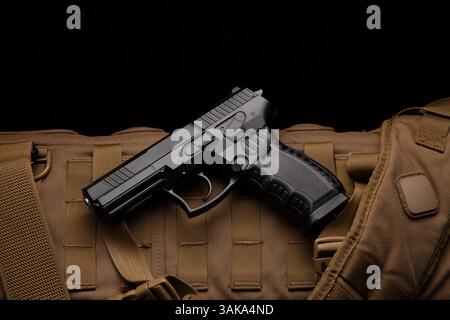 Black tactical pistol resting on brown military backpack, top-down view, dramatic contrast against dark background Stock Photo