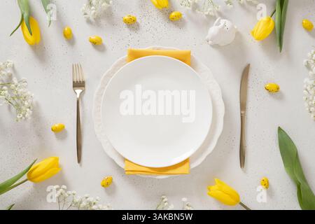 Easter table setting with spring, yellow tulips, empty white plates, bunny, eggs on white background. Happy Easter celebration. Copy space. View from Stock Photo