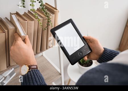 Young man with e-reader and books on shelf at home, closeup Stock Photo