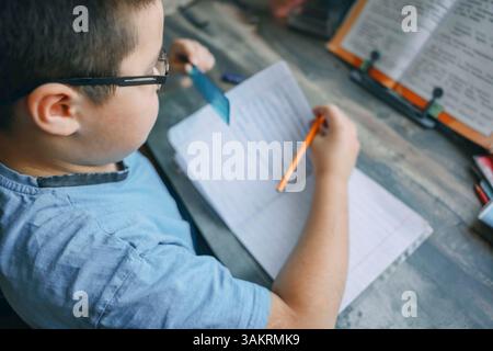 boy in blue shirt using ruler and pencil while doing homework, close up of child writing in notebook at wooden desk with textbook Stock Photo