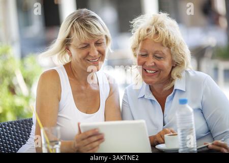Happy senior women enjoying the time in outdoor cafe. Smiling friends using touch pad and having drinks Stock Photo