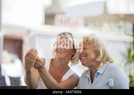 Smiling senior female friends taking selfie outdoor using smart phone. Cheerful women making happy shots, copy space above Stock Photo