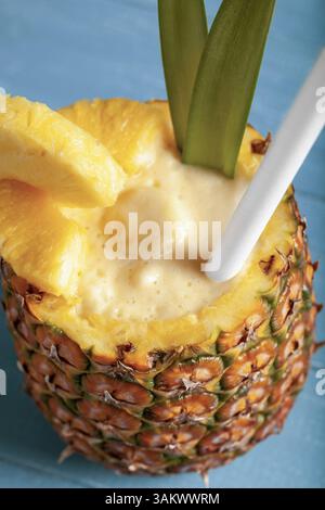 A close-up vertical photo featuring a pina colada cocktail served in a hollowed-out pineapple with pineapple wedges, a straw, and green leaves on a bl Stock Photo