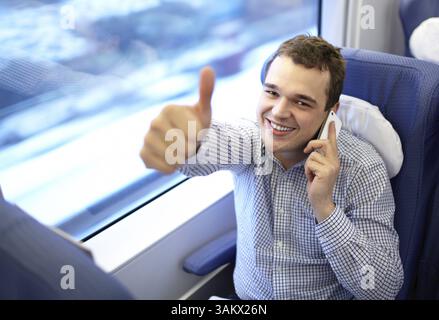 Young successful smiling businessman in modern high speed train is talking smartphone and showing his thumb up Stock Photo