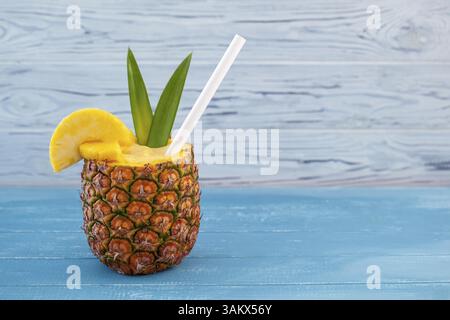 A horizontal photo featuring a pina colada cocktail served in a hollowed-out pineapple with pineapple wedges, a straw, and green leaves on a blue wood Stock Photo