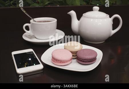Serving of a freshly brewed cup and pot of tea and a plate of macaroons with a tablet computer on a dark background Stock Photo