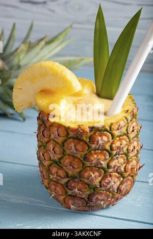 A vertical photo of a pina colada cocktail on a blue wooden background with pineapple pieces, a white straw, two green leaves, and a sliced pineapple Stock Photo