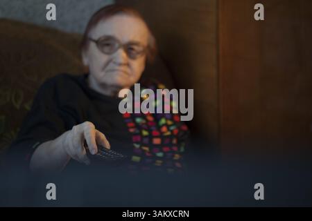 View from behind the television set of an elderly lady watching television using the remote to change channels Stock Photo
