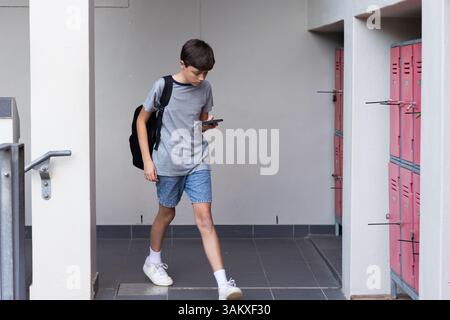 Walking in school hallway, boy with backpack using smartphone near lockers Stock Photo