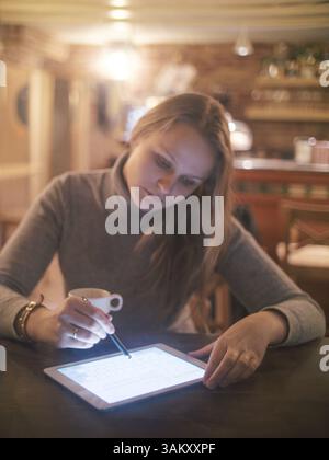 Young woman is sitting at table and typing a message on tablet PC using stylus Stock Photo