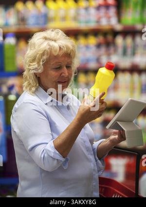 Senior woman choosing detergent in the shop. She checking the bottle label and using tablet computer to be sure with safety of goods Stock Photo