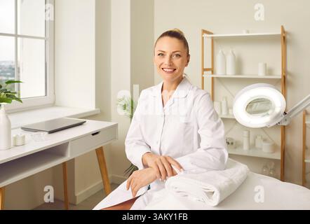 Young smiling woman cosmetologist sitting in beauty spa salon on workplace and looking at camera. Stock Photo