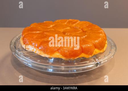 Freshly baked Tarte Tatin placed on a glass plate, showing glossy caramelized apple topping and golden pastry crust under soft indoor light Stock Photo
