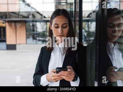 Portrait of a young female entrepreneur using a smartphone while leaning against the glass of a business building Stock Photo