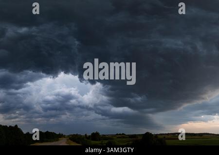 A dramatic mass of dark storm clouds descends over a quiet countryside road in Lithuania. The layered textures of the clouds convey the intensity of t Stock Photo