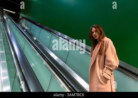 Young woman dressed with a brown coat using the escalators in the Frankfurt city subway station. Millennials woman with a serious face looking away Stock Photo