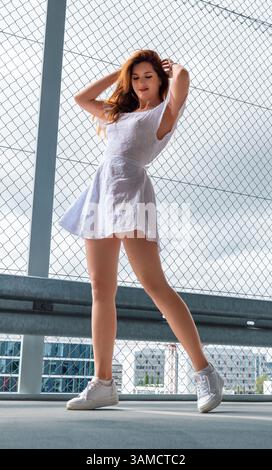 Low angle view with a beautiful millennial woman dancing in a parking lot at sunrise. Young woman wearing an elegant white dress and sneakers Stock Photo