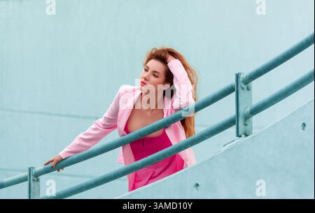 Portrait of a millennial woman in bright pink clothes leaning on a rail in a concrete walls parking lot. Young woman looking away and contemplating wh Stock Photo