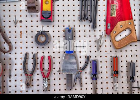 A variety of hand tools including pliers, screwdrivers, and a saw organized on a white pegboard wall in a workshop or garage setting. Stock Photo