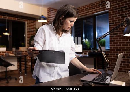 Late at night in a brick wall office, a focused businesswoman stands at her desk, comparing documents on clipboard and laptop. She carefully examines data, ensuring accuracy before making decisions. Stock Photo