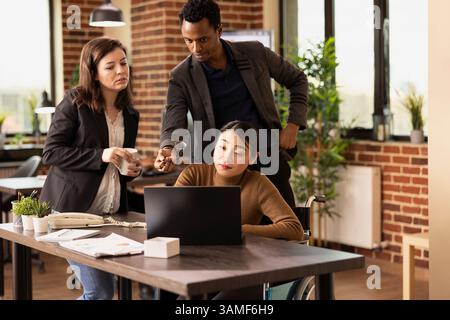 Female accountant in wheelchair analyzing business analytics with colleagues in brick wall office. Multiracial startup team surrounds a laptop on desk, collaborating to improve financial strategies. Stock Photo