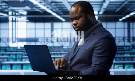Software developer in server hub checking recovery plan on laptop, monitoring hardware energy consumption. African american worker uses notebook in data center, ensuring rigs are functioning, camera A Stock Photo
