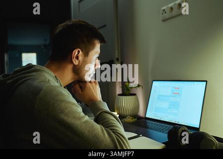 Concentrated freelancer working late at home office. Focused man in hoodie writes notes while working on laptop. Concept of night work Stock Photo