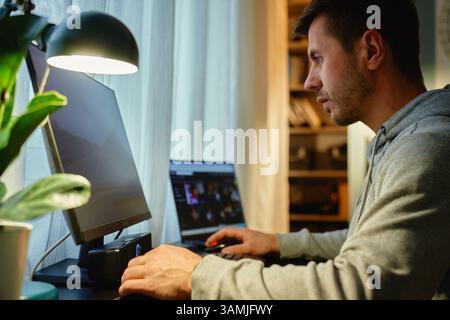 Focused man sitting by table and looking at computer monitor. Concentrated freelancer using desktop and laptop by window. Remote work from home office Stock Photo
