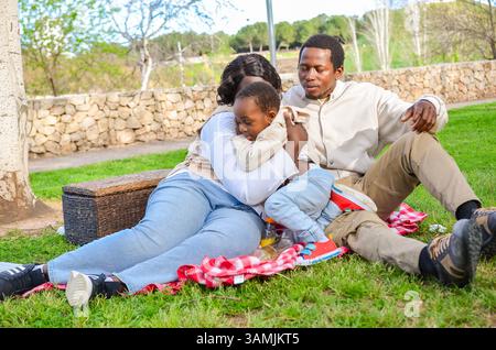 Family enjoying a relaxing picnic in a sunny park, with mother embracing son and father sitting nearby Stock Photo