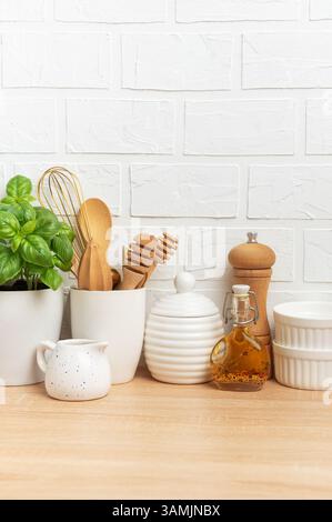 Various kitchenware, wooden utensils, fresh basil plant, ceramic ramekins, and condiments are neatly arranged on a wooden countertop against a white b Stock Photo