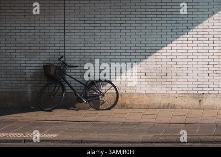 A lone bicycle with a front basket leans against a sunlit white brick wall on a quiet urban sidewalk. Stock Photo