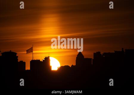 A dramatic sunset over the silhouetted skyline of Kyiv, with the sun partially obscured by buildings and a flagpole displaying the Ukrainian flag with Stock Photo