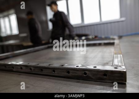 A metal frame with drilled holes sits on a workbench in a workshop, with two blurred figures of workers in the background near windows. Stock Photo
