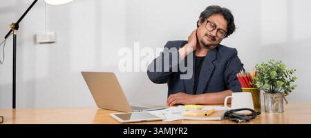 A tired, uncomfortable Asian businessman sits at his desk in front of a computer, suffering from office syndrome, massaging his neck and shoulder to r Stock Photo