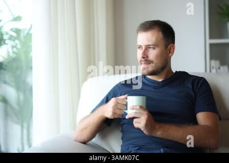 Confident man drinking and looking through a window sitting on a couch at home Stock Photo